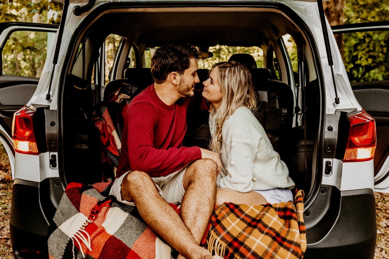 A couple sits in the trunk of their SUV in a fall forest.