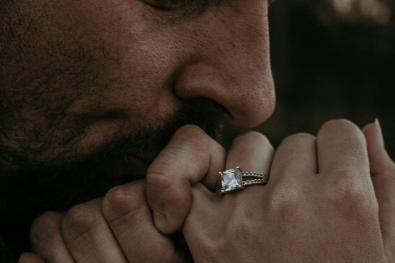 a man links pinkies with his lover and kisses her hand, with a split shank princess cut ring.