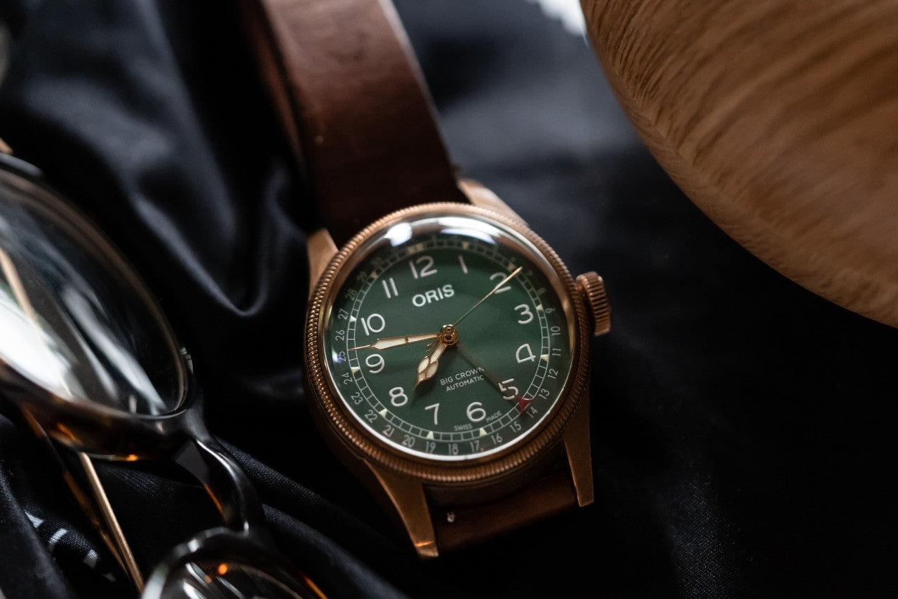 A close-up of an Oris watch displayed on a wooden surface next to a pair of sunglasses.