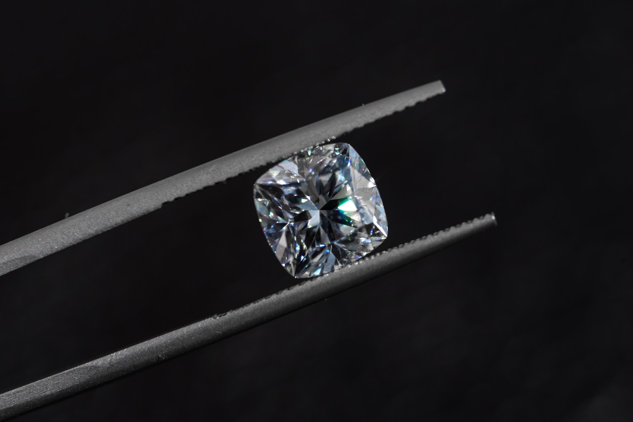 A close-up of a cushion cut diamond held between the prongs of jeweler’s tweezers.