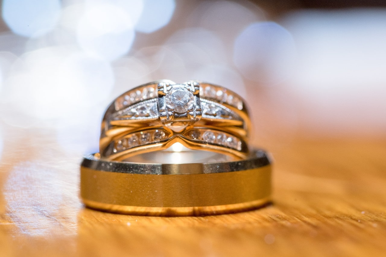 A close-up of a woman’s wedding band and engagement ring stacked atop a men’s wedding band on a wooden surface.