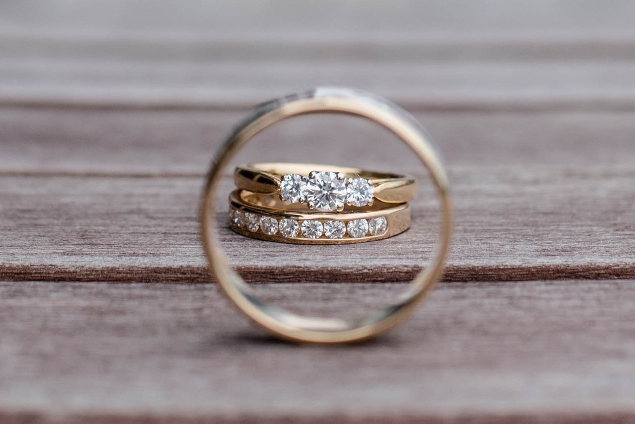 A matching engagement ring and wedding band seen through a men’s wedding band on a wooden surface.