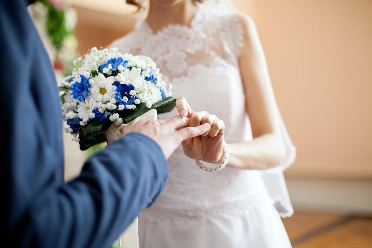 A close-up of a bride and groom at the altar, the bride placing a wedding band on her groom’s finger.